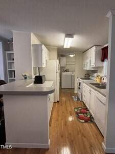 A galley kitchen with white cabinets, white appliances, and wood flooring; a laundry area with a washer and dryer is visible at the far end. There are floral rugs and a small countertop with a toaster.