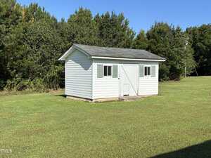 A small white shed with gray roof, green shutters, and a double door sits on a grassy lawn with trees and bushes in the background under a clear blue sky.