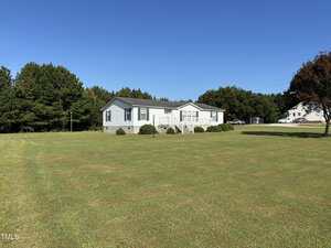A single-story, light blue house with white trim sits on a large, well-maintained grassy lawn with a few trees and bushes. The sky is clear and blue, and another house is visible in the background.