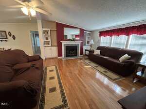 A living room with hardwood floors, two brown sofas, a fireplace with a red accent wall, a large mirror above the mantel, two small area rugs, and windows with red valances letting in natural light.