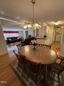 A dining area with a wooden table and chairs sits on a rug, next to a kitchen with white cabinets. In the background is a living room with a brown sofa, armchair, coffee table, and large window with red curtains.