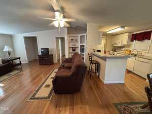 A living room with hardwood floors, brown couches, a TV on a wooden stand, and a kitchen with white cabinets and appliances. A breakfast bar with two stools separates the kitchen from the living area.