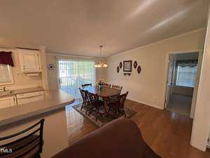 A dining area with a wooden table and six chairs is set near a sliding glass door leading outside. A chandelier hangs above the table. To the left is a partial view of a kitchen, and a doorway leads to another room.