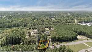 Aerial view of a wooded area with a highlighted rectangular property near a road, surrounded by trees and scattered houses under a blue sky with light clouds.