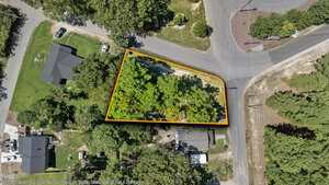 A triangular, tree-covered vacant lot is bordered by roads on three sides in a residential area, highlighted with an orange outline. Nearby, several houses and driveways are visible through the surrounding trees.