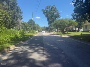 A paved residential street with a white car parked in the distance, flanked by grass, trees, power lines, and houses under a clear blue sky. Shadows from trees are cast onto the road.