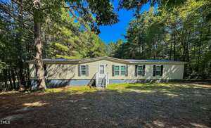 A beige, single-story manufactured home with a small front porch and steps, set in a wooded area with tall trees and dappled sunlight on the ground.