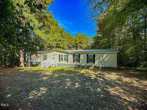A single-story manufactured home with beige siding and green shutters sits on a spacious, shaded lot surrounded by tall trees. A small white-railed porch with steps leads to the front door.