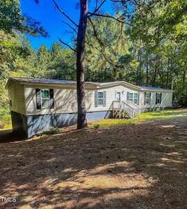 A beige manufactured home with white trim sits on a raised foundation in a wooded area. Sunlight filters through tall trees, and a small white staircase leads to the front door. The ground is covered with pine needles.