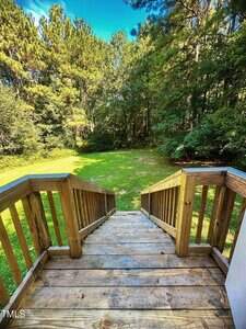 View from the top of a wooden staircase leading down to a grassy backyard bordered by dense trees under a bright blue sky.