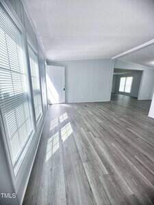 A bright room with light gray walls, large windows with blinds, light wood flooring, and a white front door. Sunlight streams through the windows, casting shadows on the floor. The space is open and unfurnished.