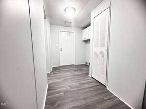 A hallway with light-colored walls and dark wood-style flooring leads to a white door. There is a louvered closet door on the right and overhead cabinets next to the door. A ceiling light fixture is centrally located.