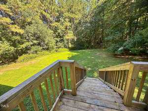 View from the top of a wooden staircase leading down to a large, grassy backyard surrounded by dense trees and greenery. Sunlight casts shadows on the lawn and trees.