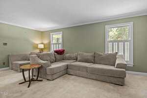 A living room with light green walls, beige carpet, a large beige sectional sofa, a small round wooden side table, a lamp, and two windows letting in natural light.