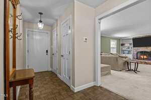 A hallway with beige walls and parquet flooring has a small wooden table and coat rack on the left. Three white doors are visible, and an open doorway leads to a living room with a fireplace and beige sectional sofa.
