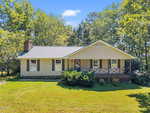 Charming single-story beige house with dark shutters and a covered front porch at 209 W Dogwood Drive in Creedmoor, set on a green lawn with bushes and surrounded by tall trees under a blue sky.