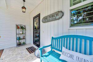 A covered porch with white siding features a black door, a teal bench with a "Welcome Friends" pillow, a patterned rug, hanging lantern, metal shelf with plants, and a sign reading "THE PORCH Sit Relax Gossip.