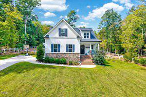 Charming two-story home at 2493 Golden Forest Drive, Franklinton, with white siding, stone accents, a spacious green lawn, trees, white fence, basketball hoop on the drive, and inviting landscaping under a partly cloudy sky.