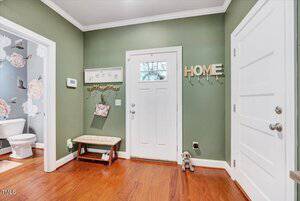 A green-walled entryway with a white front door, a small bench, a wall hook rack with bags, and "HOME" letter hooks. A bathroom with floral wallpaper is visible through an open door on the left.