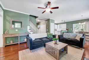 A living room with green walls, a ceiling fan, two sofas, a wooden coffee table, and a sideboard. The space opens to a kitchen and dining area with hardwood floors and natural light from large windows.