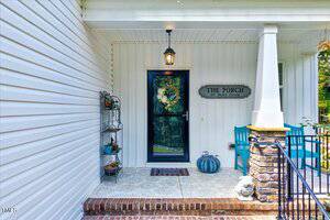 A covered porch with a black-framed glass door, a welcome mat, a shelf with plants, a blue chair and table, a light fixture, and a decorative sign reading "The Porch Sit Relax Gossip" on a white wall.