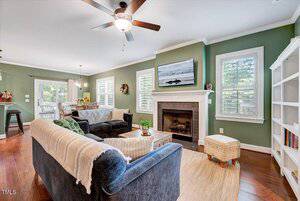 A living room with green walls, hardwood floors, a ceiling fan, a gray sofa and loveseat with blankets, a fireplace with a TV above it, two windows, a bookshelf, and a dining area visible in the background.