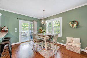A dining area with green walls, a wooden table set for four, a flower arrangement centerpiece, a small rug, and a bench with pillows under a window. Glass doors lead to an outdoor space with a view of greenery.