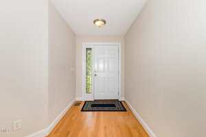 A hallway with light-colored walls and wood flooring leading to a white front door with a glass panel on the left side. A small black and beige rug is placed in front of the door.