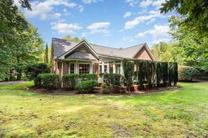 A single-story brick house with tall trimmed shrubs along the side, surrounded by grass, trees, and a partly cloudy sky in the background. The house has large windows and a gabled roof.
