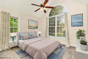 A bedroom with a large window, beige walls, a gray upholstered bed with mauve bedding, two nightstands with lamps, plants, a blue patterned rug, floral curtains, and ocean-themed wall art. Ceiling fan overhead.