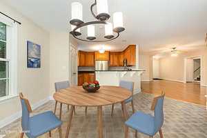 A dining area with a round wooden table, four gray chairs, a bowl of fruit, and a modern chandelier. The space opens to a kitchen with wood cabinets and stainless steel appliances and a living area with light wood flooring.