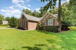 A one-story brick house with white trim and a large arched window sits on a well-maintained lawn, bordered by bushes and trees under a partly cloudy sky. A concrete driveway is visible on the left.