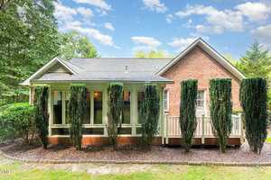 A single-story brick house with a gable roof, large windows, and a row of tall, narrow evergreen trees in front. A small porch with white railings is visible to the right, surrounded by a landscaped yard.