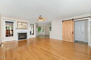 Spacious living room with hardwood floors, a ceiling fan, fireplace with white mantel, large windows, and a wooden sliding barn door leading to another room; open floor plan with view into kitchen area.