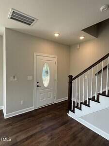 A white front door with an oval window is beside a wooden staircase with white risers and dark railings. The entryway has gray walls, dark wood flooring, and recessed ceiling lights.
