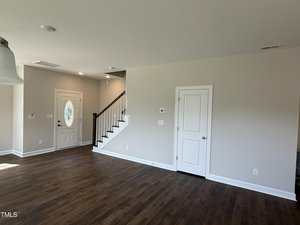 A room with dark wood flooring, beige walls, a white front door with an oval window, a white interior door, and a staircase with a dark handrail and white balusters. Ceiling lights and vents are visible.