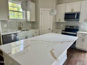 Modern kitchen with white cabinets, stainless steel appliances, marble-patterned island countertop, gold faucet, two pendant lights, and a window above the sink letting in natural light.