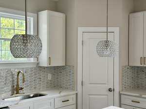 A modern kitchen with white cabinets, gold hardware, hexagonal tile backsplash, a gold faucet, and two pendant lights with circular crystal designs. A window is above the sink and a closed white door is visible.