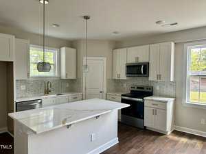 A modern kitchen with white cabinets, marble countertop island, stainless steel appliances, two pendant lights, tile backsplash, and windows letting in natural light. Dark wood flooring contrasts with the light-colored decor.