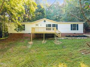 Charming single-story house with white siding and black shutters on a brick foundation at 3122 Willow Creek Drive, Wake Forest. A wooden deck with railing leads to the front door, surrounded by trees and a grassy front yard.