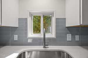 A kitchen sink with a silver faucet is centered beneath a small window. The backsplash is light gray tile, and there are two electrical outlets on each side. Trees are visible through the window.