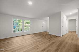 A spacious, unfurnished room with light-colored walls, wood flooring, large front windows, and a white door. A hallway leads to other rooms, and natural light brightens the space.