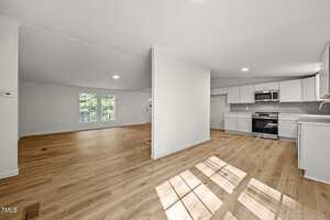 Open-plan interior with light wood floors, white walls, and a partial dividing wall. The space includes a kitchen with white cabinets and stainless steel appliances, and a living area with large windows.