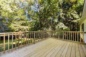 A wooden deck with a simple railing overlooks a green backyard bordered by tall trees and dense foliage on a sunny day.