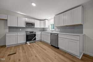 Modern kitchen with white cabinets, stainless steel appliances, gray subway tile backsplash, light wood flooring, and recessed lighting. A window is above the sink, and there are no visible items on the counters.