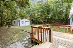 A wooden deck attached to a house overlooks a grassy backyard with a small concrete path leading to a white metal garage. The yard is bordered by dense trees and forest under clear daylight.