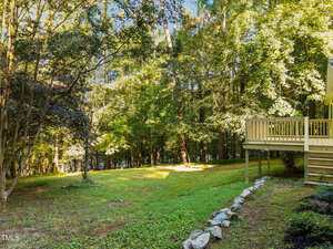 A grassy backyard bordered by a line of rocks and mature trees, with a wooden deck attached to the right side of a house. Sunlight filters through the trees, creating areas of light and shadow on the lawn.