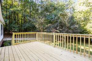 A wooden deck with a simple railing overlooks a green backyard with grass and mature trees. The deck is attached to a house on the left, and sunlight filters through the surrounding forested area.