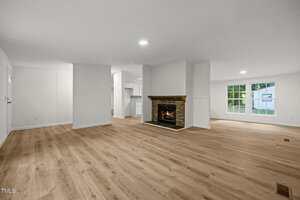 Spacious open-concept living area with light wood flooring, white walls, a stone fireplace in the center, and large windows letting in natural light. The kitchen is visible in the background.