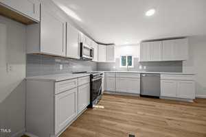 A modern kitchen with white cabinets, stainless steel appliances, gray tile backsplash, light wood flooring, and a small window above the sink. The space is well-lit with recessed ceiling lights.